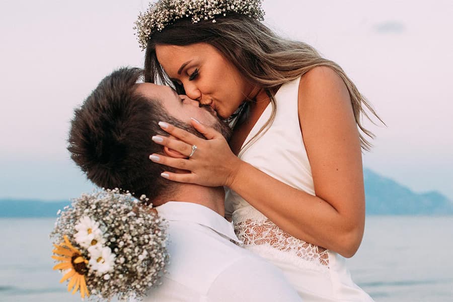 A couple shares a romantic kiss on a beach at sunset, with the woman wearing a floral crown.
