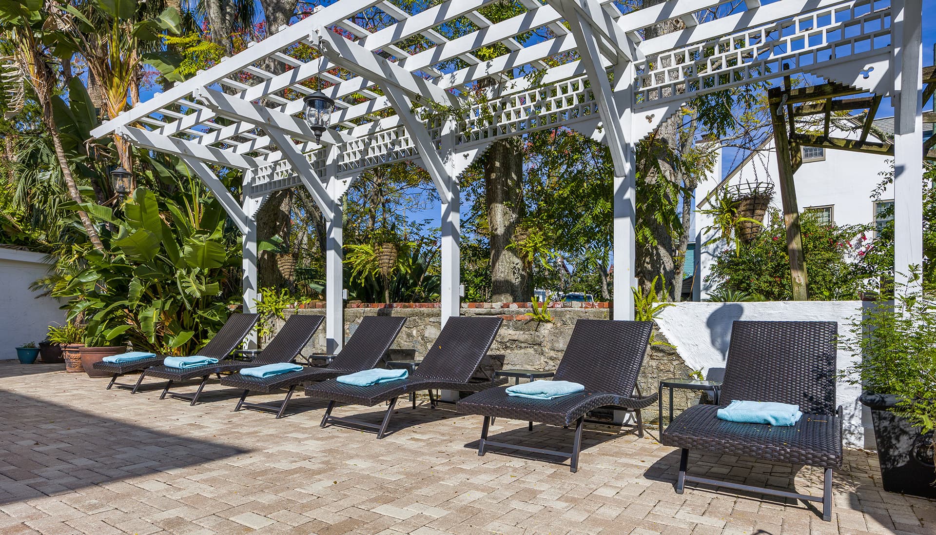 A row of black lounge chairs with blue cushions under a white pergola surrounded by greenery.