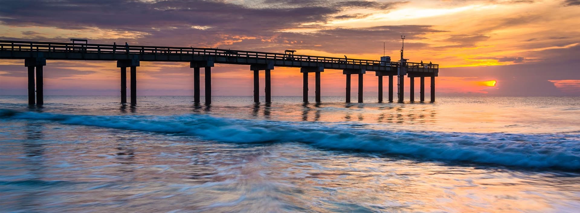 A wooden pier extends over a calm ocean at sunset, with vibrant colors reflected in the water.