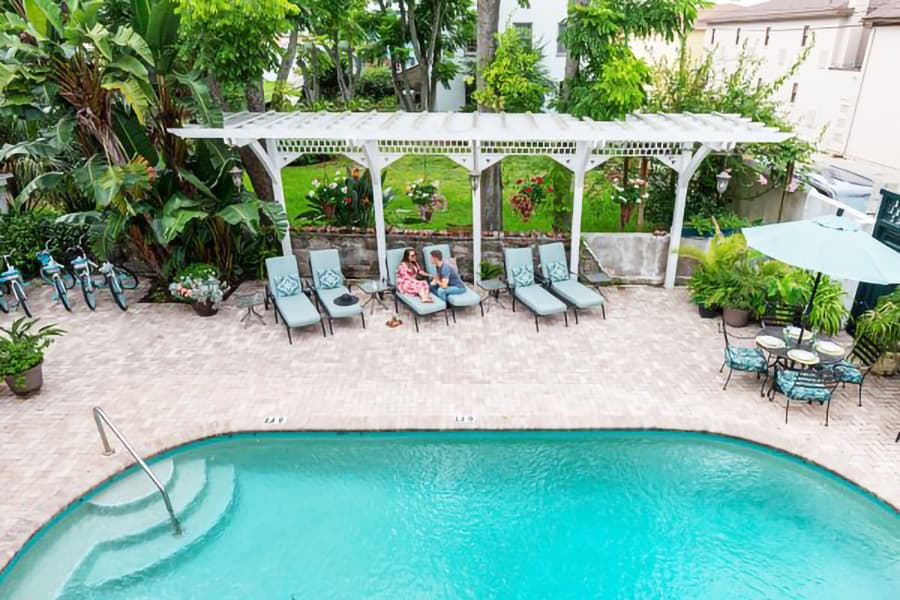 A poolside area featuring lounge chairs, lush greenery, and two people relaxing under a pergola.