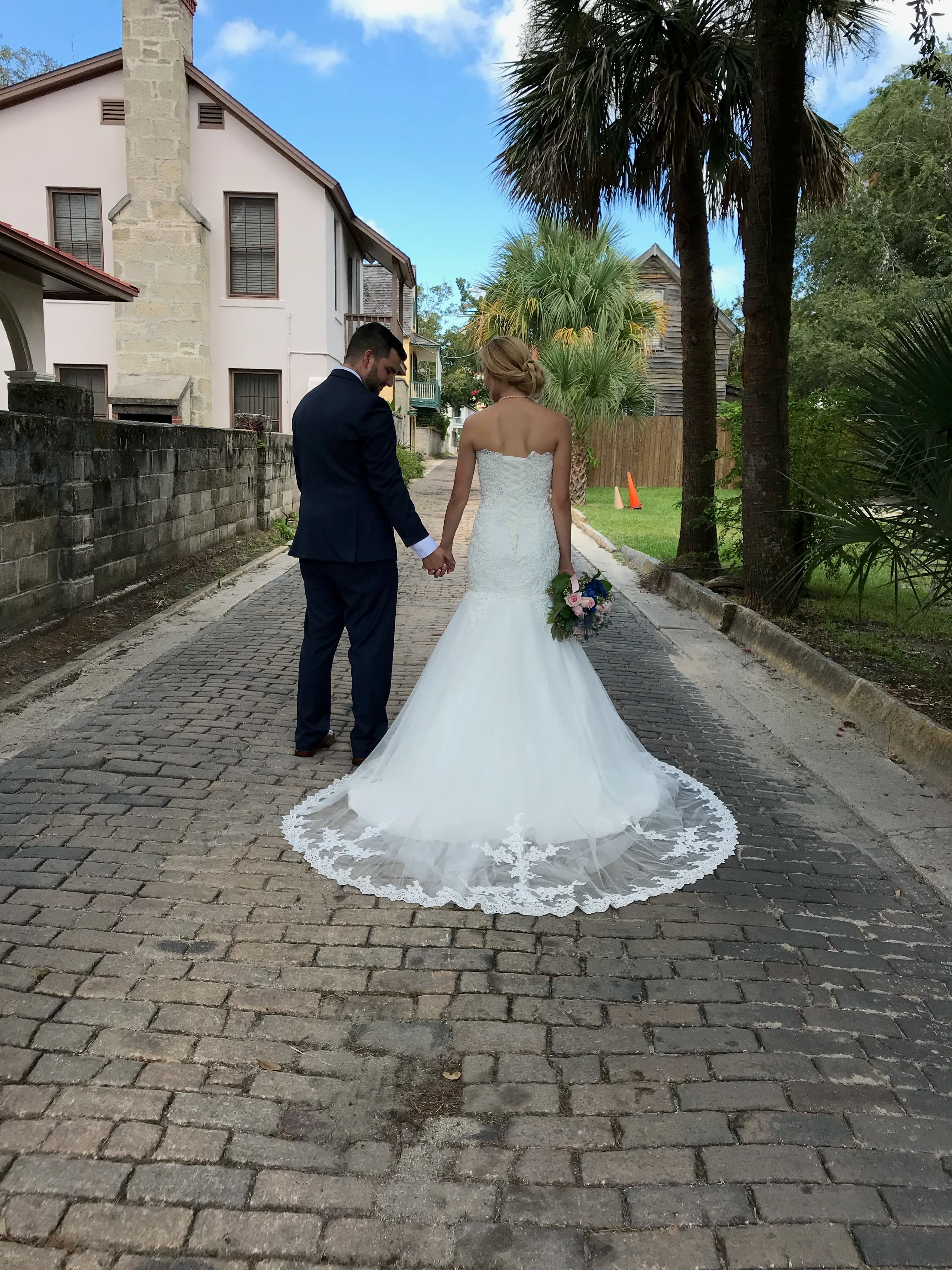 A couple holds hands while walking down a cobblestone path, surrounded by greenery and historic buildings.