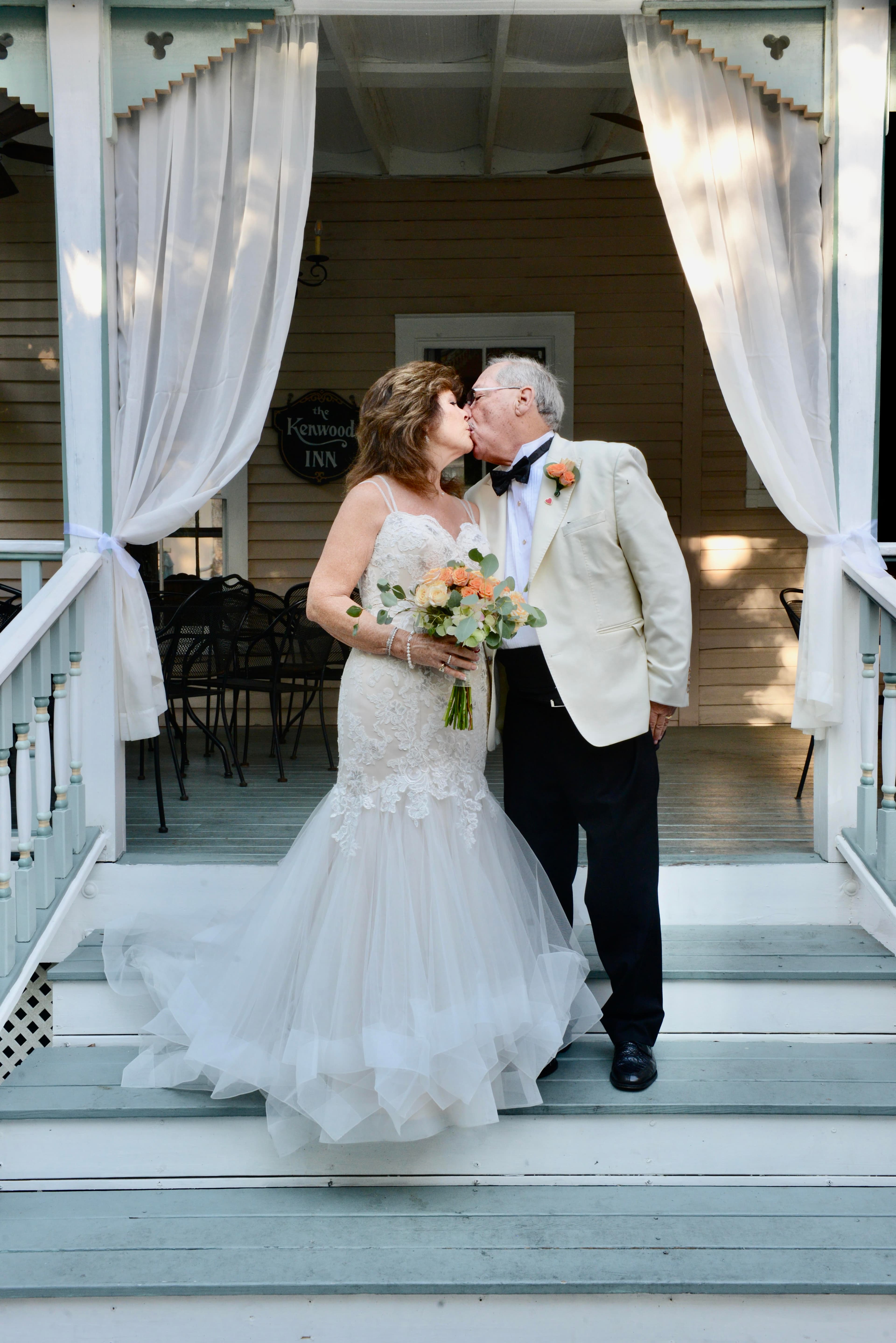 A couple in formal wedding attire shares a kiss on the steps of a charming inn, holding a bouquet of flowers.