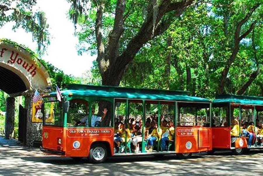 A bright orange trolley filled with passengers is parked under lush green trees near an archway labeled "Fountain of Youth."