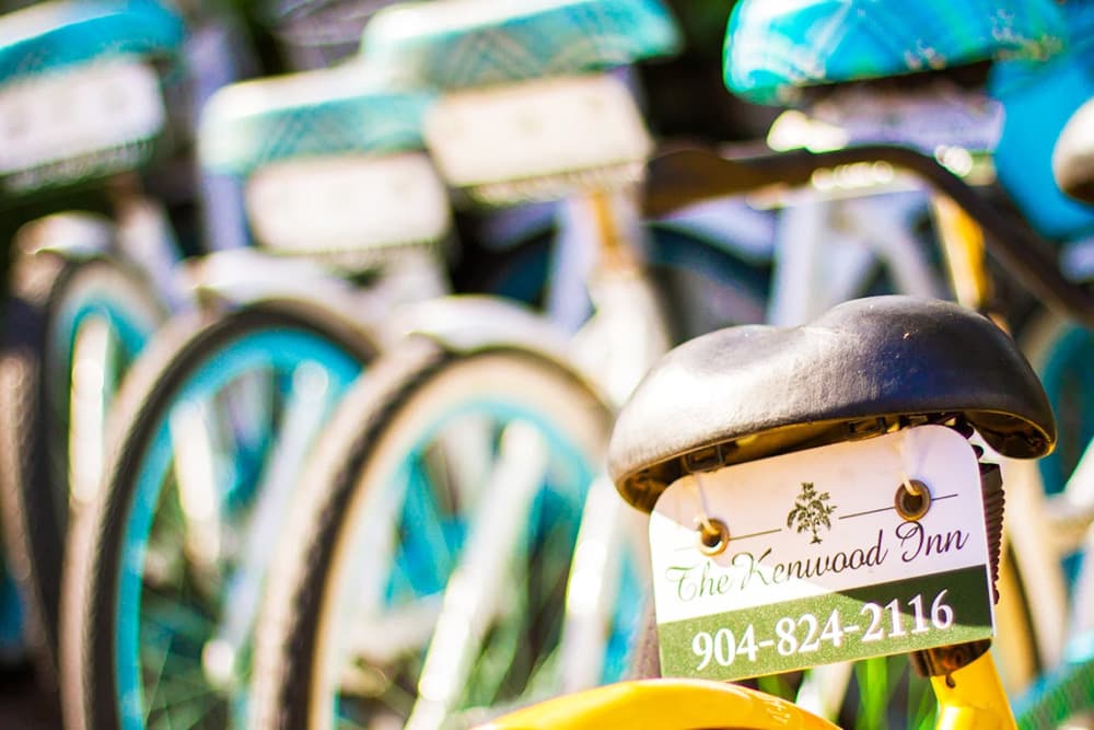 A close-up of a bike's saddle featuring a tag that reads "The Kenwood Inn" along with the phone number 904-824-2116, with blue and white bicycles blurred in the background.