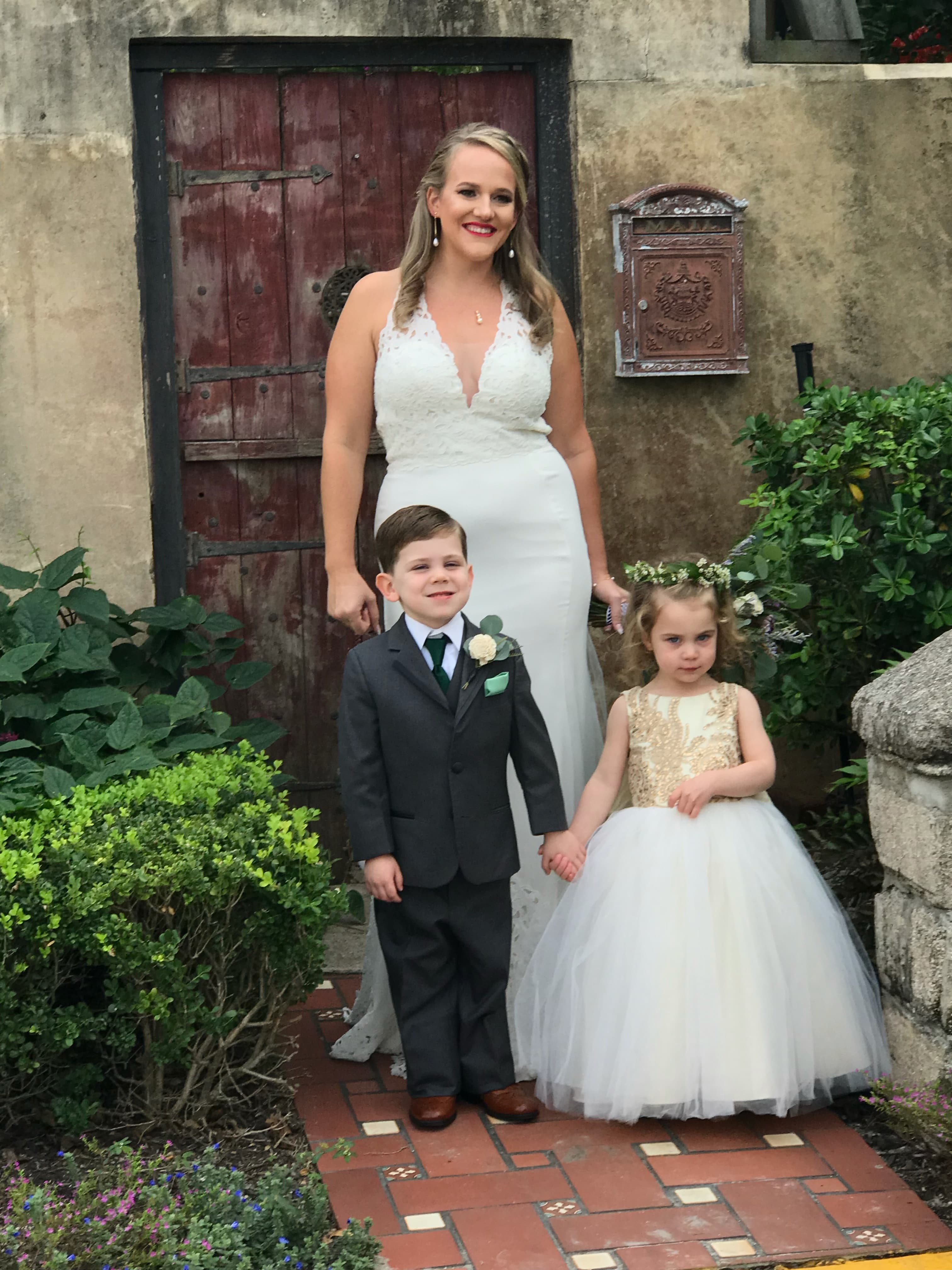 A smiling woman in a white gown stands with a boy in a suit and a girl in a white dress, outdoors near a rustic door and greenery.